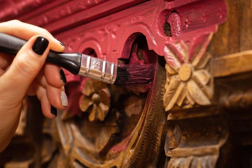 Egy személy közelről mély rózsaszín festékkel fest egy díszes, faragott fafelületet ecsettel. A bonyolult faragások virágmotívumokat tartalmaznak, és a friss festék alatt még látható az eredeti fa színe. A személy keze, fekete és ezüst körömlakkal, óvatosan viszi fel a festéket a részletes mintázatra.