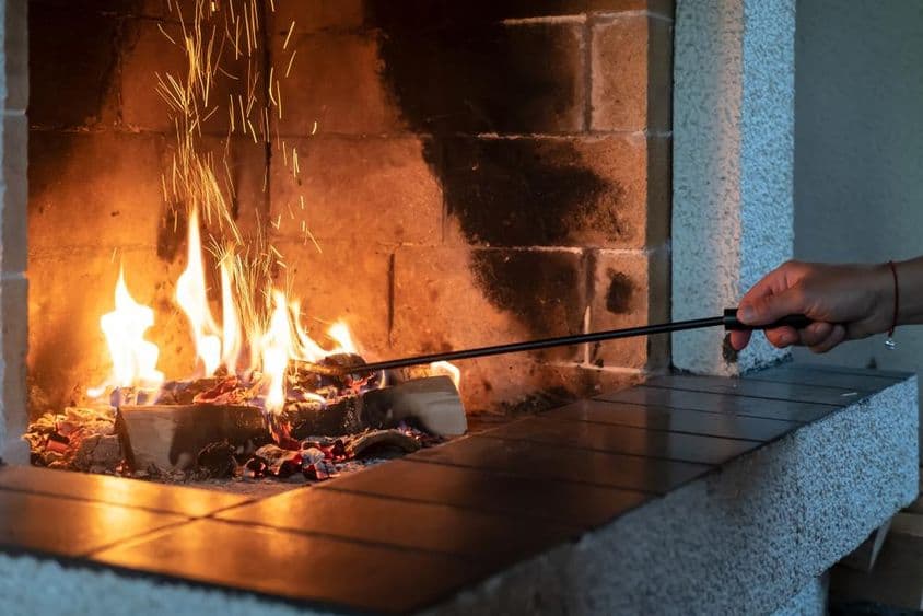 A női kéz vas piszkálóval egyenesíti ki az égő tűzifát egy nyitott kandallóban