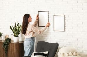 Fiatal nő üres képkeretet akaszt fehér téglafalra a nappaliban.