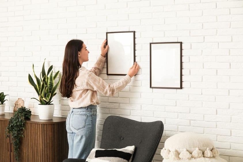 Fiatal nő üres képkeretet akaszt fehér téglafalra a nappaliban.
