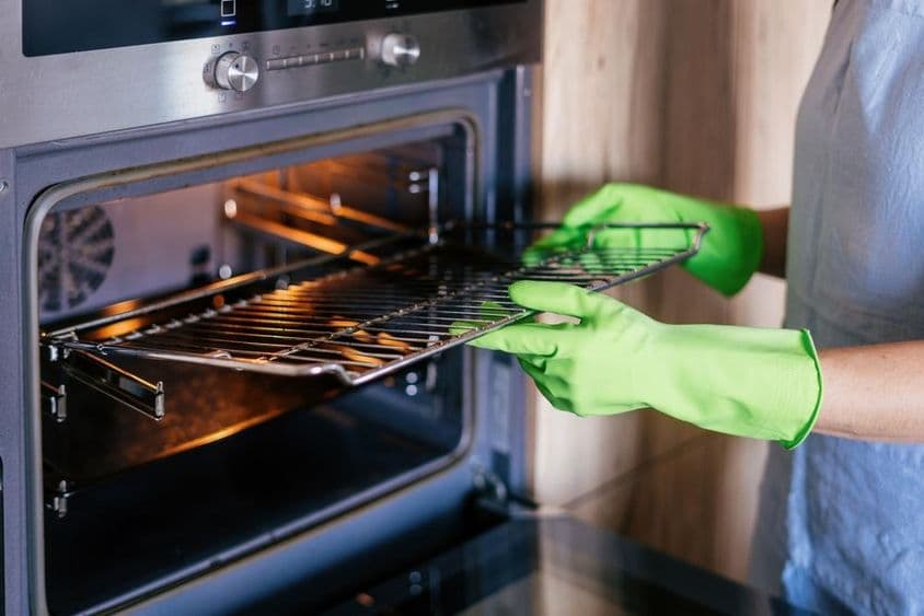 Egy zöld tisztítókesztyűt viselő nő óvatosan eltávolít egy piszkos rácsot a modern sütőből.